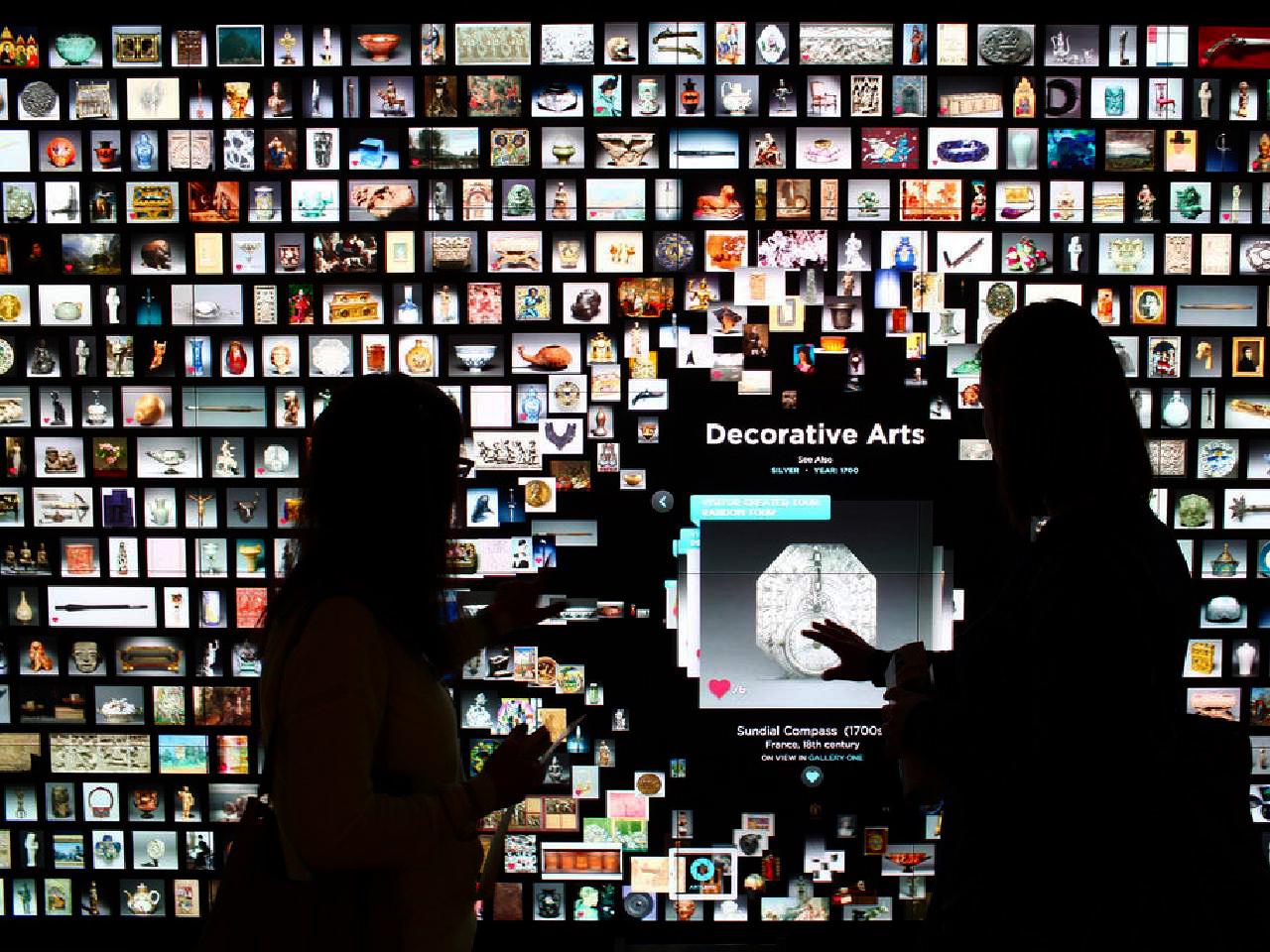 Image of two people in front of a large screen with hundreds of images of works of art. Both are touching the screen and immediately between the two figures a larger artwork image is visible and the words 'decorative arts' are immediately underneath it.