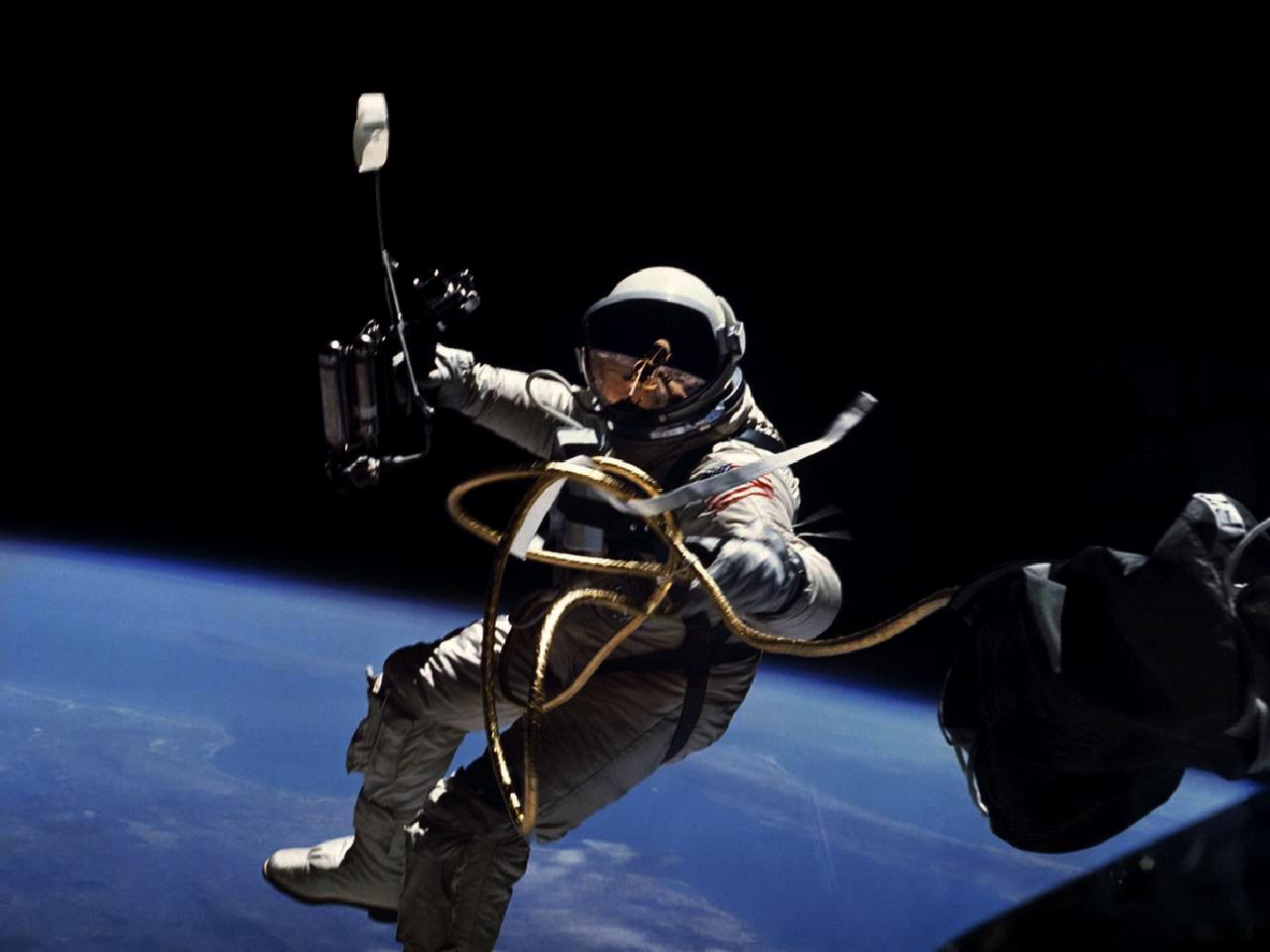 An astronaut in a white spacesuit is shown floating with the Earth's horizon in the background. The astronaut is connected to an unseen spacecraft via an umbilical, which extends into the foreground.