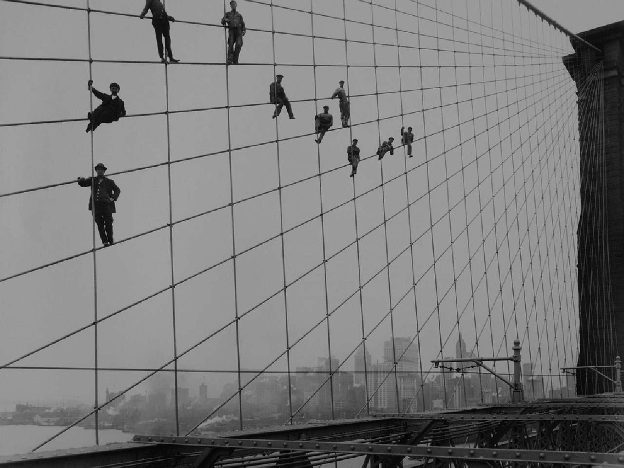 Early photograph of eight men perched on supporting cables on the Brooklyn Bridge during its construction.