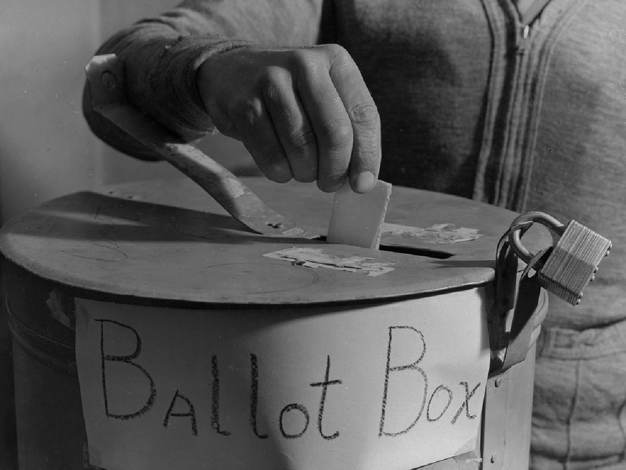 Close-up of a hand dropping a paper ballot into a cylindrical metal ballot box with a slot in the top. The box is locked with a padlock. On the front of the box are the words 'ballot box.'