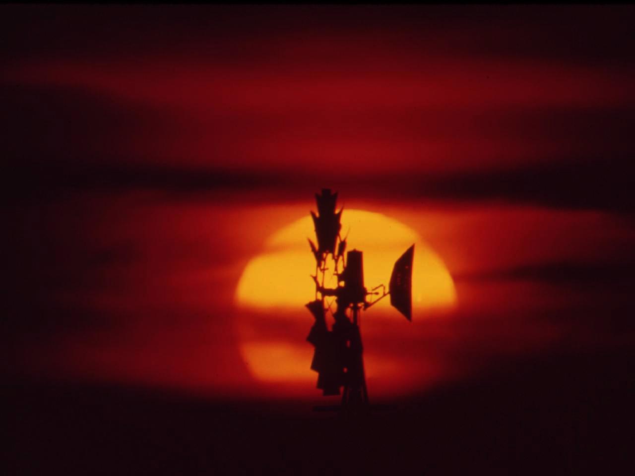 A farm windmill is silhouetted against an orange-red sunset.