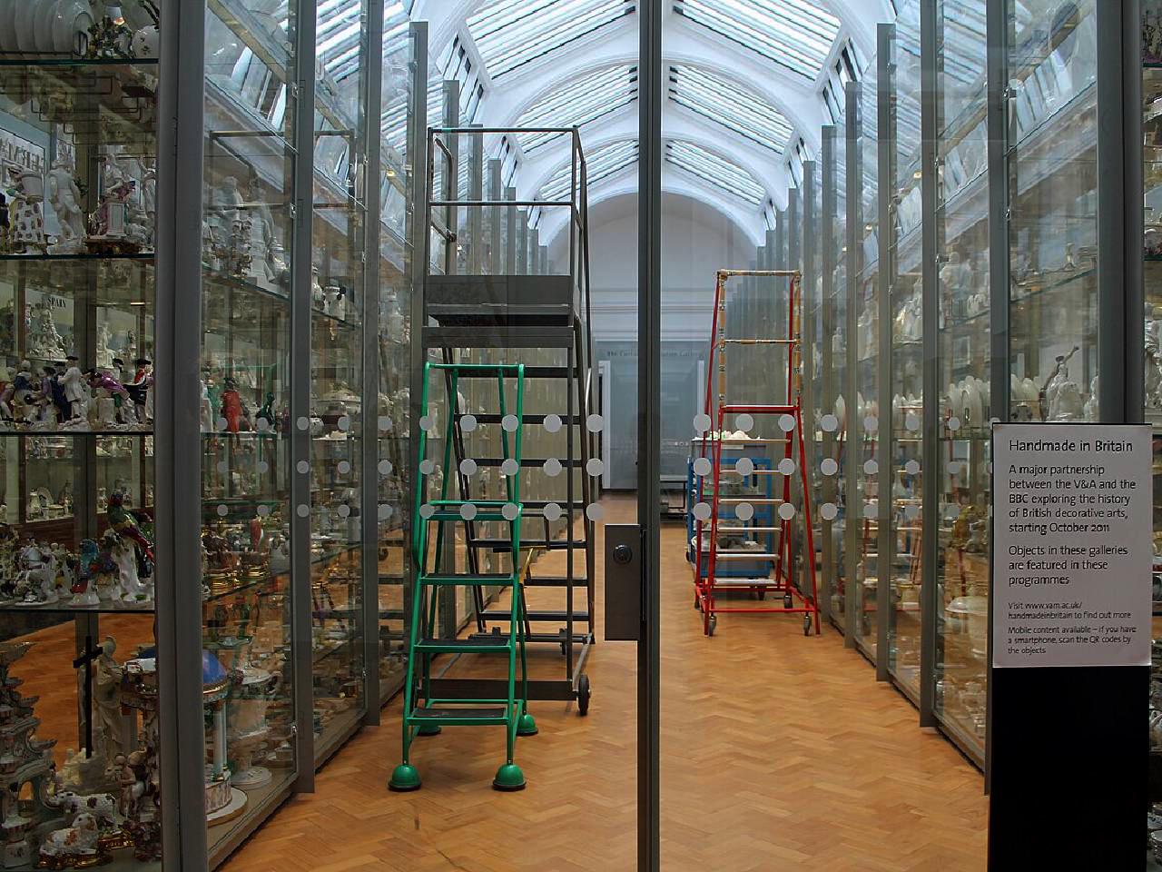 Glass storage cases full of decorative arts on the left and right side of a narrow walkway, with green and red movable staircases.