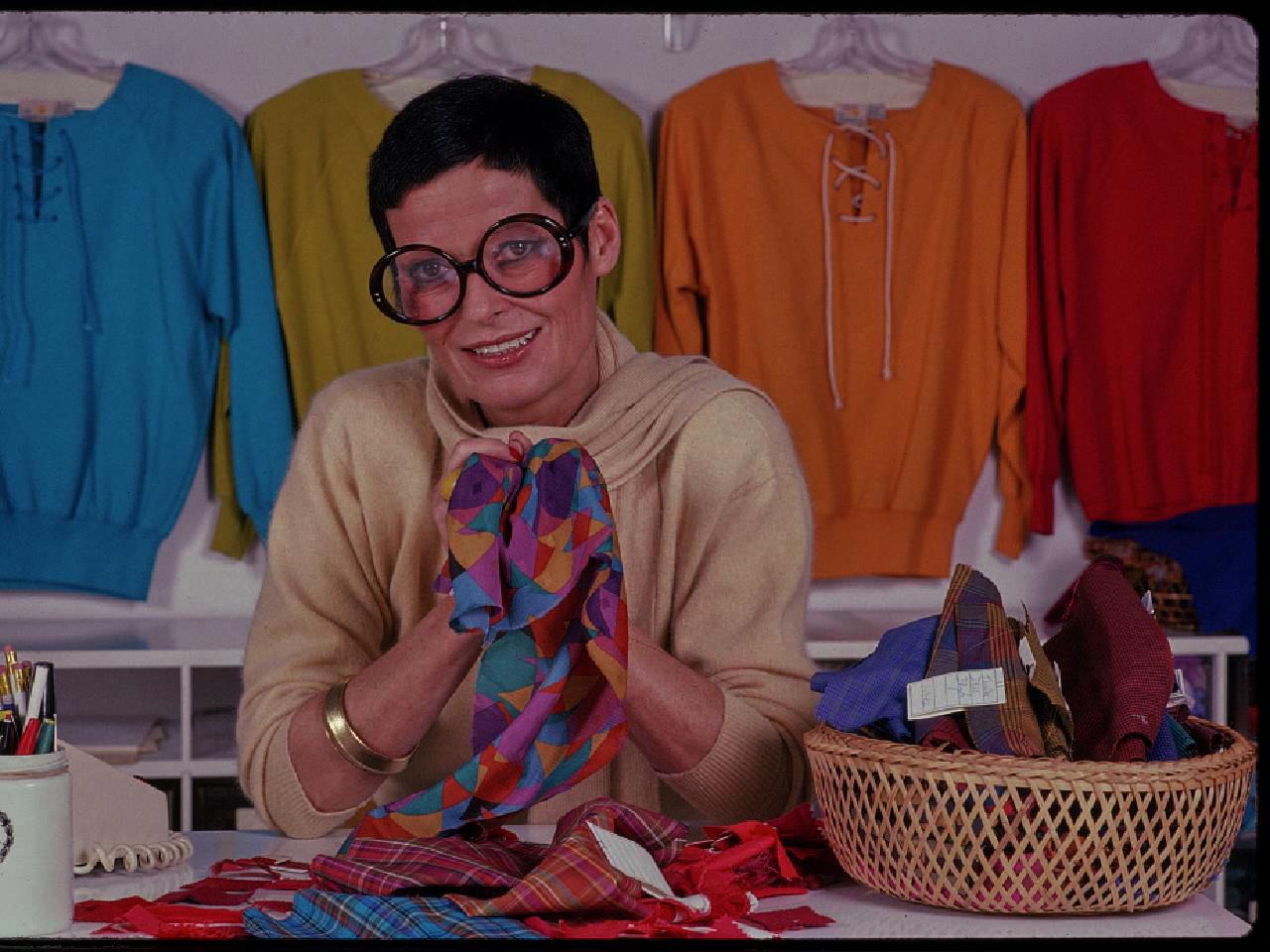 Color image of a woman with short black hair and large round black glasses seated at a table with many colorful bolts of fabric in her hands, to either side of her, and behind her.