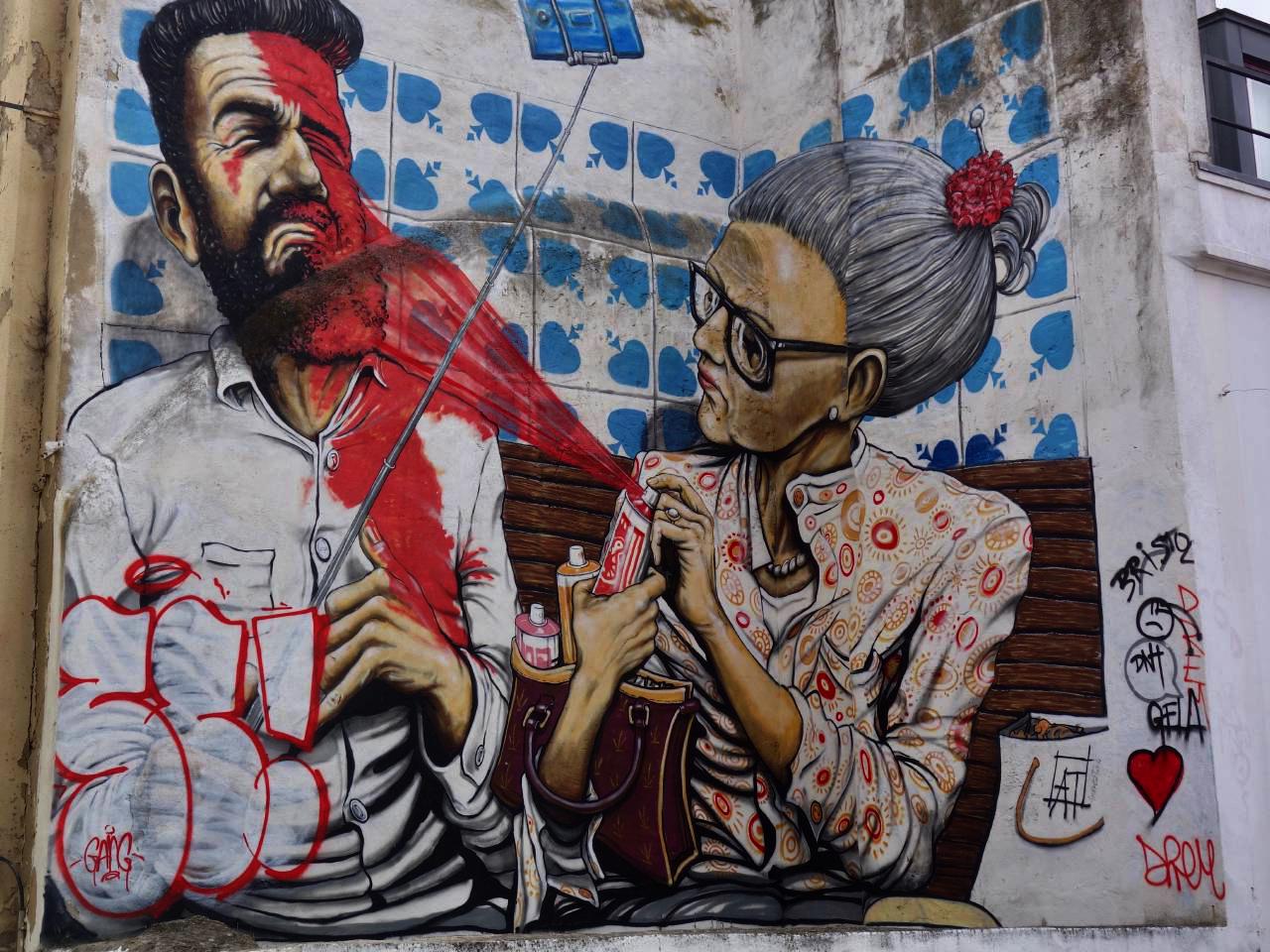 Graffiti on a building wall depicting an older woman spraying a younger man in the face with red spray paint. The man is holding a selfie stick with a phone attached.