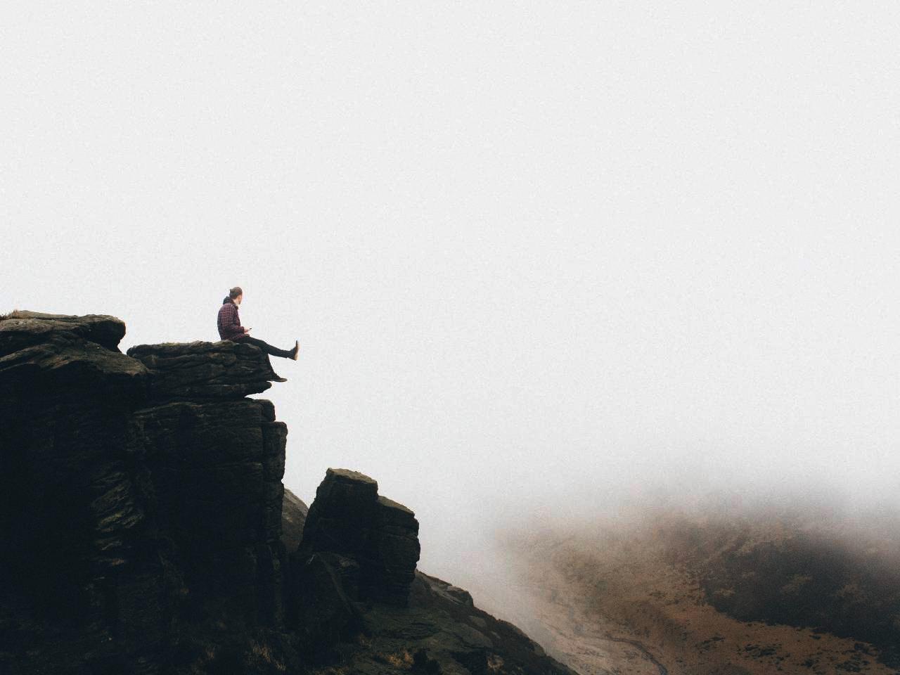 Image of a man sitting on the edge of a rock-face cliff. He is kicking his feet up and fog is directly beneath him.