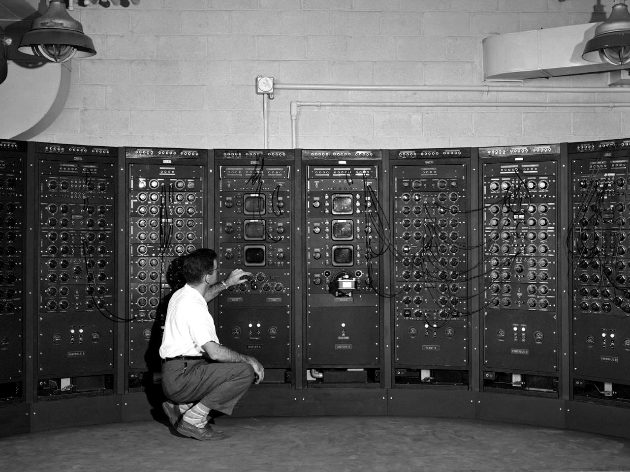 Black and white image of a man crouched in front of a bank of analog computers, with numerous switches, readouts, and cables.